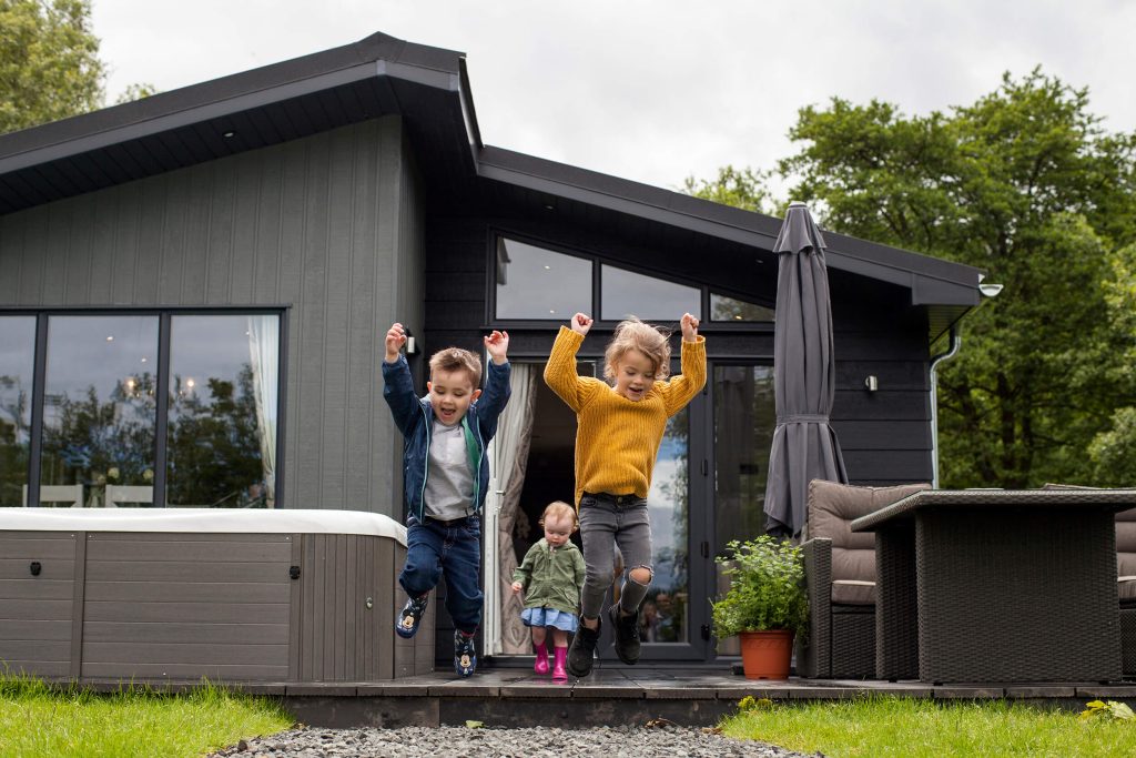 Three young children on a family friendly stay at Kielder Waterside. Two of the children are jumping together from the front step of their hot tub lodge, whilst the third runs up behind them.